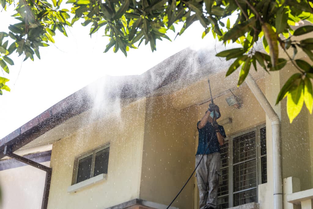A person power washes the exterior of a two-story home. The individual is wearing protective eyewear and a blue shirt with grey trousers.