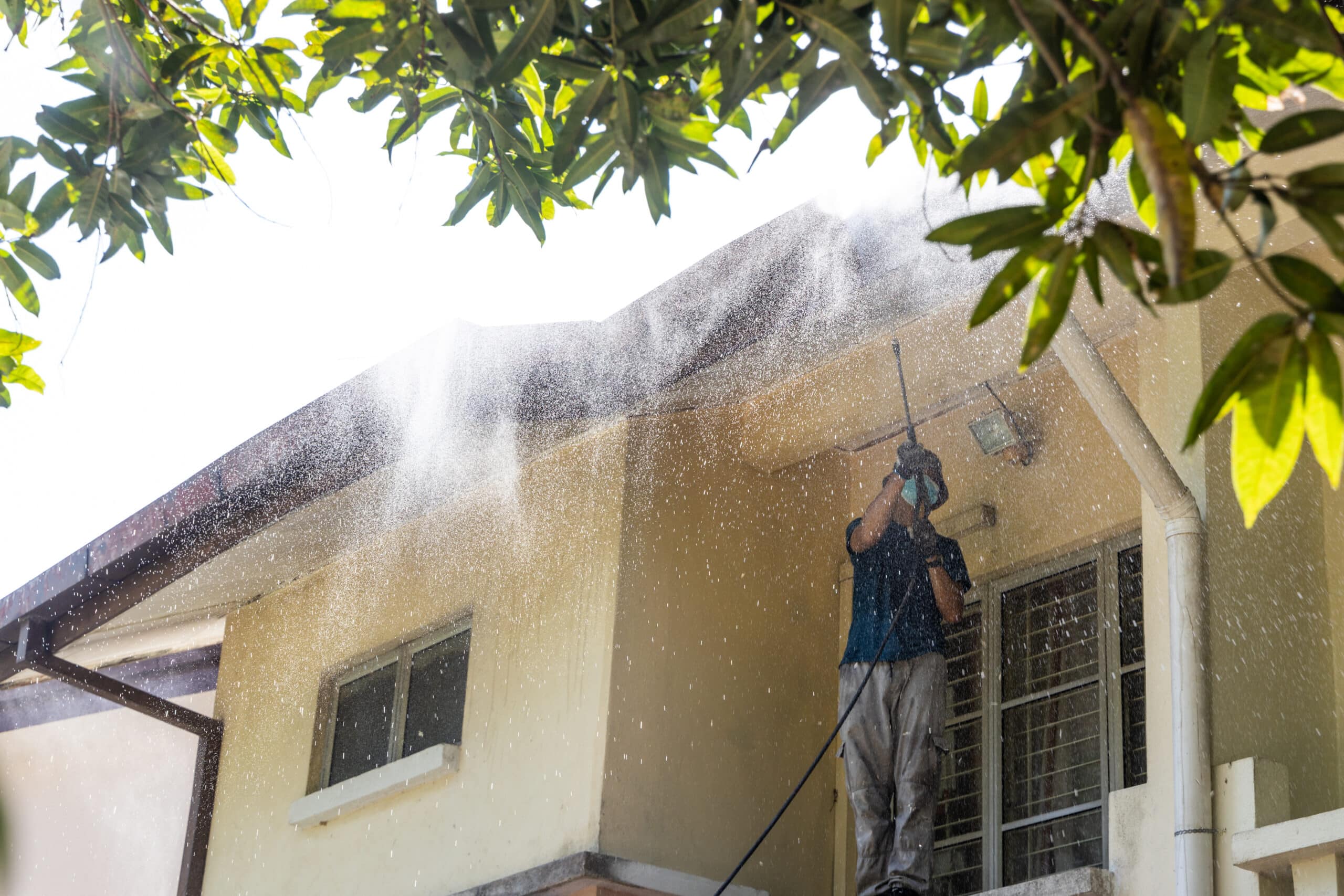 A person power washes the exterior of a two-story home. The individual is wearing protective eyewear and a blue shirt with grey trousers.