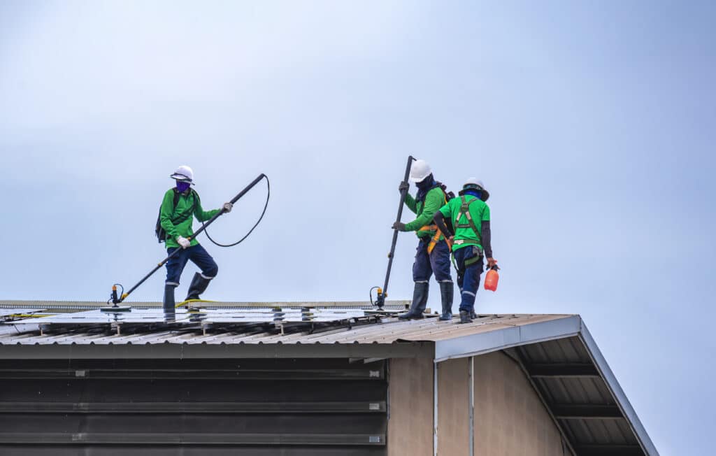 Three workers wearing hard hats, safety vests and green tops stand on the roof of a commercial building with solar panels for a roof cleaning service in Roanoke, VA.